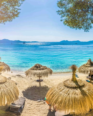Tropical beach with thatched umbrellas, loungers, turquoise water, sailboats, and forested islands under clear blue sky