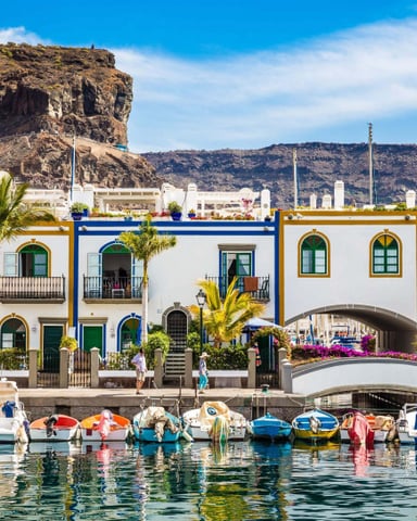 Colorful waterfront buildings with green shutters and palm trees, moored fishing boats in harbor, rocky cliff backdrop under blue sky
