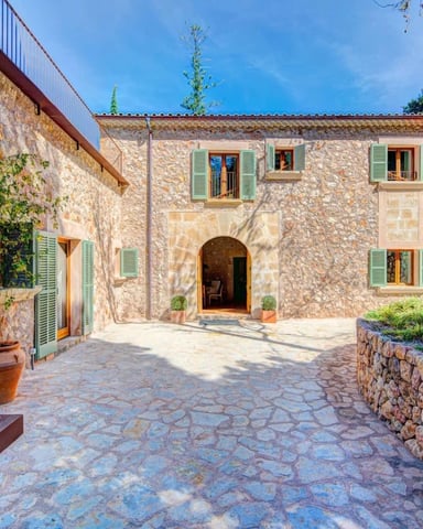 Stone villa courtyard with arched entrance, green shutters, and paved terrace surrounded by trees