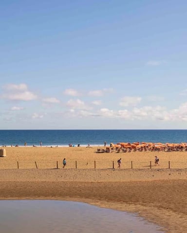 Sandy beach with shallow tidal pool in foreground, rope barriers, scattered beachgoers, and calm blue ocean under partly cloudy sky