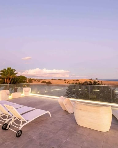 Rooftop terrace with lounge chairs, modern seating, palm trees, and panoramic sea and countryside views at dusk
