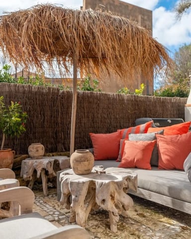 Outdoor patio with wooden furniture, orange and gray cushions, driftwood tables, thatched palm umbrella, and fireplace.