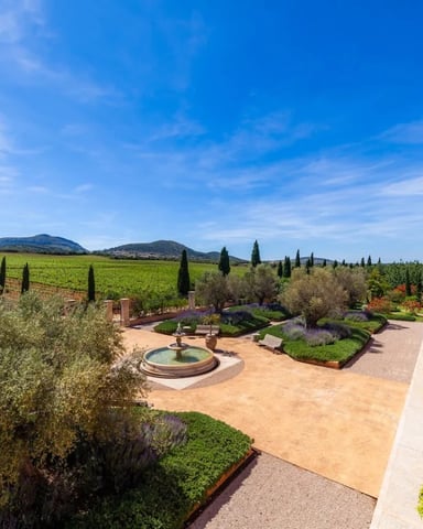 Tuscan villa courtyard with fountain, manicured gardens, and vineyard-covered hills under blue sky