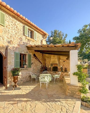 Stone villa courtyard with white metal chairs, pergola, arched doorway, and Mediterranean landscaping under clear blue sky