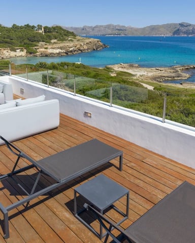 Wooden terrace with white lounge seating and sunbeds overlooking turquoise Mediterranean bay with rocky coastline and forested hills