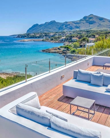 Rooftop terrace with white sofas overlooking turquoise Mediterranean bay, mountain, and coastal village