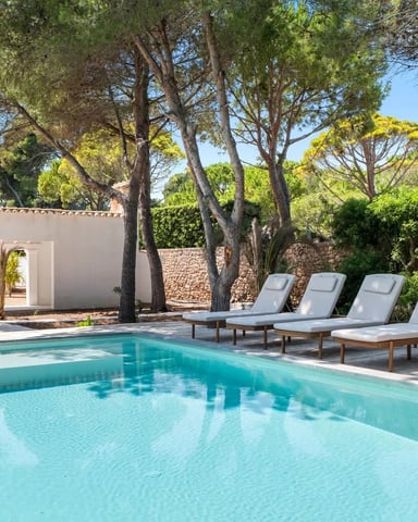 Swimming pool with turquoise water, four white lounge chairs, white modern house, and tall pine trees under clear blue sky