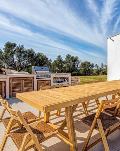 Outdoor patio with wooden dining table, woven chairs, built-in grill, and white house wall overlooking green lawn and trees