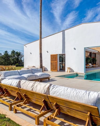 Modern white villa with pool, wooden lounge chairs with cushions on patio, dining area, and palm tree against blue sky
