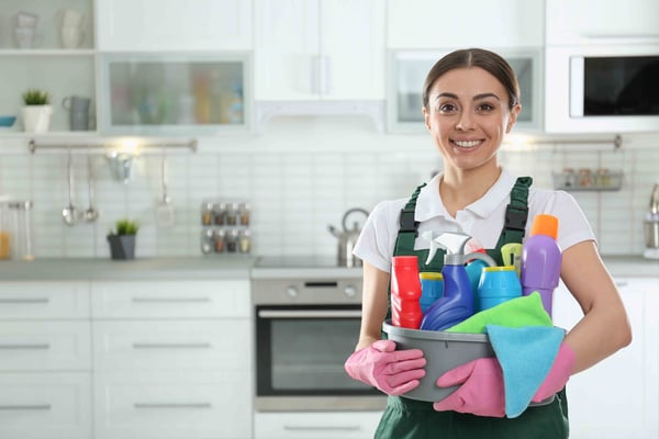 Smiling woman in green apron holding caddy of cleaning supplies in modern white kitchen