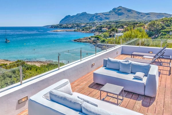 Rooftop terrace with white sofas overlooking turquoise Mediterranean bay, mountain, and coastal village