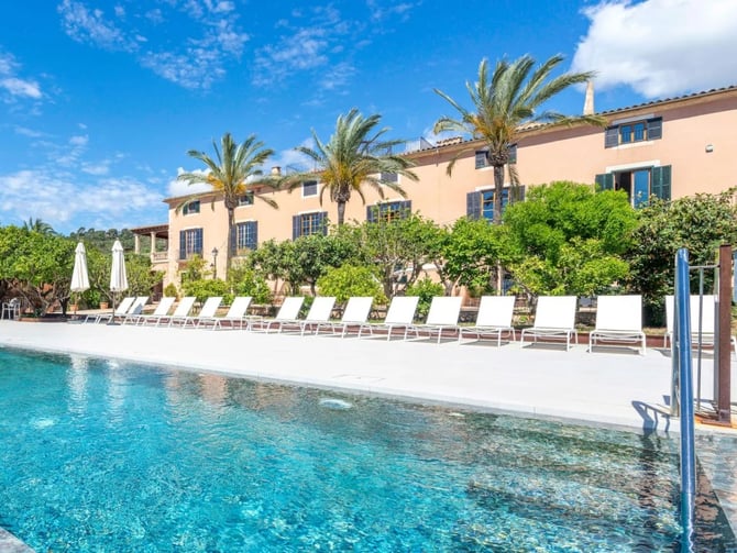 Swimming pool with white loungers and umbrellas facing peach-colored villa with palm trees and blue shutters